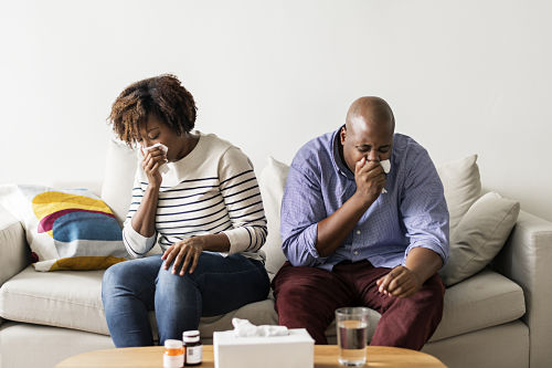 sick young couple on couch holding tissues to noses